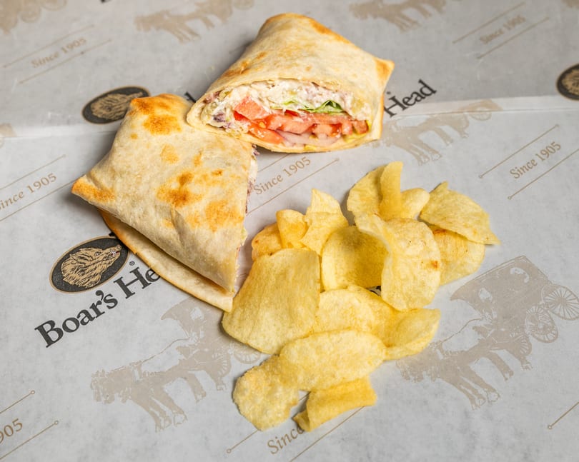 Boar's Head wrap, cut in half, filled with meat, cheese, and vegetables, served with potato chips.