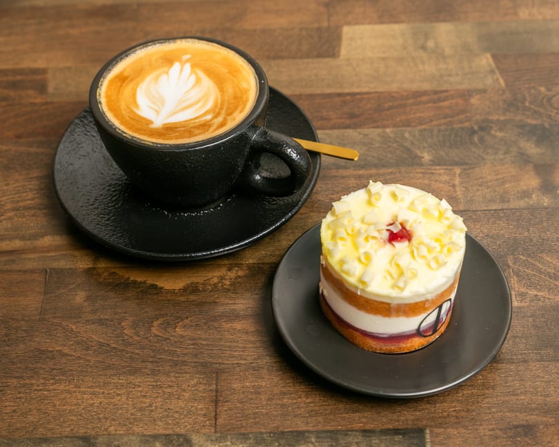 Latte with latte art and a layered cake with white frosting and chocolate curls on a wooden table.
